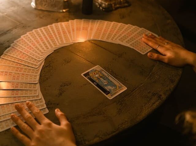 Foto de cartas do Tarot, formando um leque e uma meia lua, com uma carta central. O tarot está em cima de uma mesa de madeira escura e existem mãos em cima das cartas e da mesa. A foto ilustra o artigo Origem e História do Tarot do site Ori Mystyco.