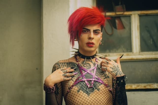 Foto de um homem andrógino e não binário com cabelos vermelhos olhando para a câmera. Ele tem cabelos vermelhos e tatuagens em formato de V do pescoço ao peito. A foto ilustra o artigo A coragem de ser você mesmo do site Ori Mystyco.