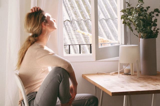 Foto de uma mulher loira, com blusa creme e calca cinza, sentade em uma cadeira, em frente a uma mesa, olhando com ar de melancolia pela janela ensolarada de um apartamento. A foto ilustra o artigo Momentos difícies são chamados para o despertar do site Ori Mystyco.