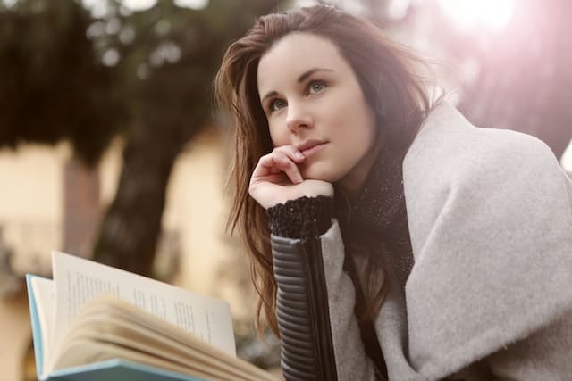 Foto de uma mulher sentada em um parque, com um livro na mão, com a outra mão no queixo e com cara de pensativa. A foto ilustra o artigo O nosso corpo somatiza os nossos pensamentos do site Ori Mystyco.