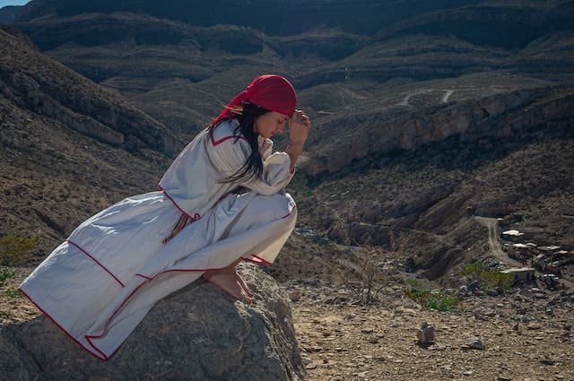 Foto de uma indígena Mexicana, com roupa branca e bordados vermelhos. Ela têm um pano vermelho na cabeça e está de cócoras orando e em cima de uma pedra no meio do deserto. A foto ilustra o artigo Oração Nahuatl - Perdão, carinho, desapego e libertação do site Ori Mystyco.