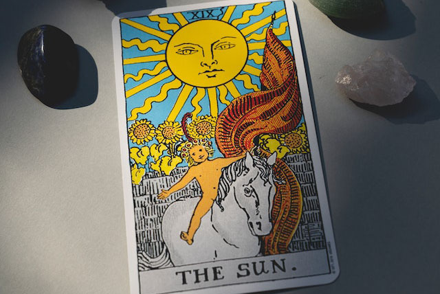Imagem da Carta O Sol do tarot Rider Waite em cima de uma mesa rodeada por cristais. A imagem serve para ilustrar o artigo Carta do Dia no Tarot de Rider Waite do site Ori Mystyco