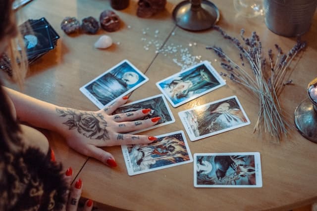 Foto de cartas do tarot em uma mesa de madeira clara, e aparece a mão de uma mulher com a mão sob uma delas. A foto ilustra o artigo Seu Tarot do Dia no Tarot de Rider Waite do site Ori Mystyco.
