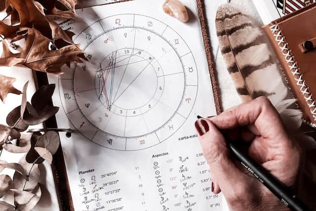 Imagem de uma mão de mulher com um lápis na mão escrevendo em um mapa astral impresso. A imagem é usada para ilustrar o artigo "As 12 Casas do Zodíaco: O que elas significam" do site Ori Mystyco