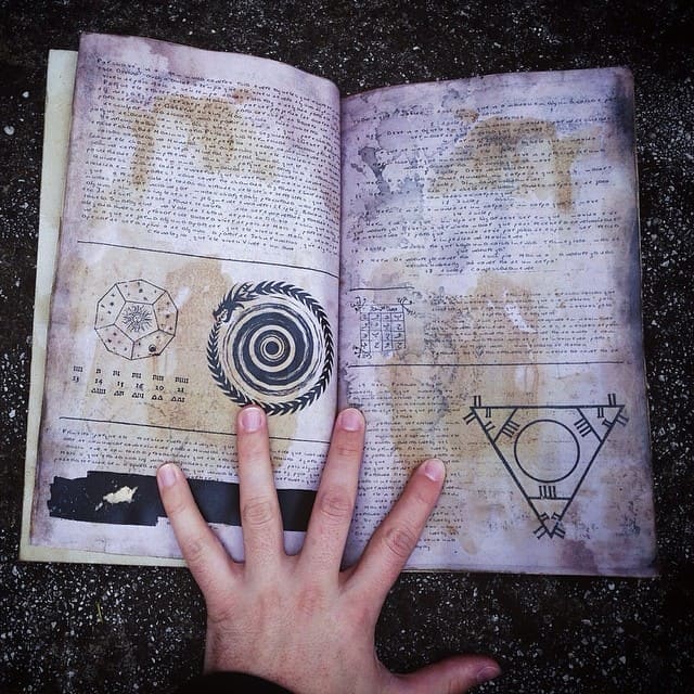 Foto com mãos de uma pessoa em cima de um livro de magia aberta, onde podemos ver textos e sigilos escritos. A foto serve para ilustrar o artigo O que são sigilos? Como eles fortalecem suas magias? do site Ori Mystyco.