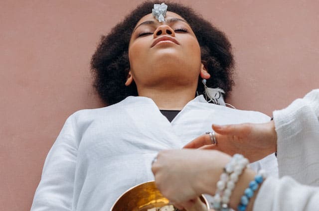 Foto de uma mulher preta deitada, com um cristal no seu chakra frontal e com um sino tibetano no seu chakra umbilical. Há a mão de outra pessoa, com pulseiras de cristais, tocando o sino tibetano. A foto serve para ilustrar o artigo O que são Chakras? Como equilibrá-los? do site Ori Mystyco.