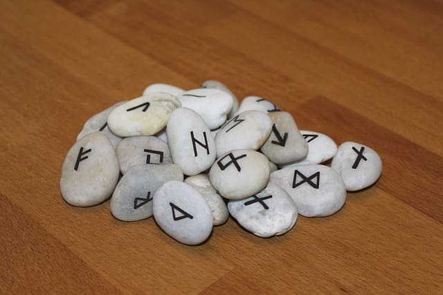 foto de runas feitas com pedra branca e letras pretas, sob uma mesa de madeira clara. A foto ilustra a página Runas Online Grátis Passado Presente e Futuro do site Ori Mystyco.
